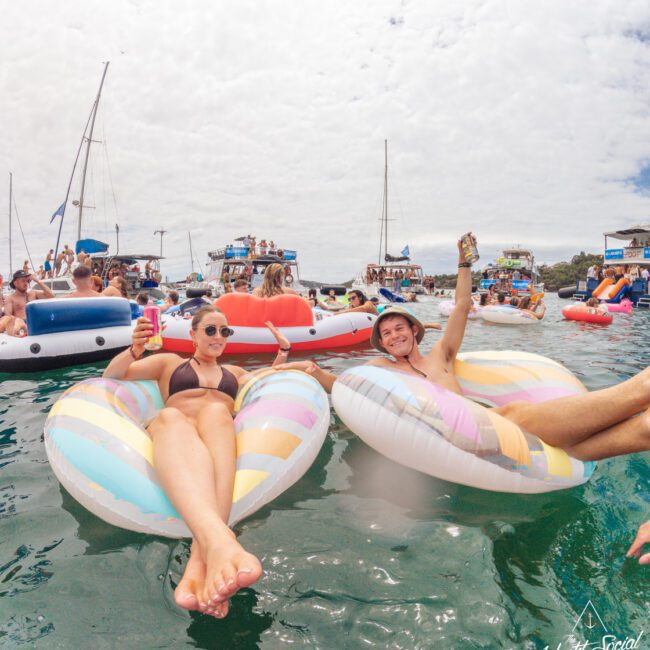 Two people relax on colorful pool floats, holding drinks and smiling, surrounded by boats and other people enjoying a sunny day on the water.