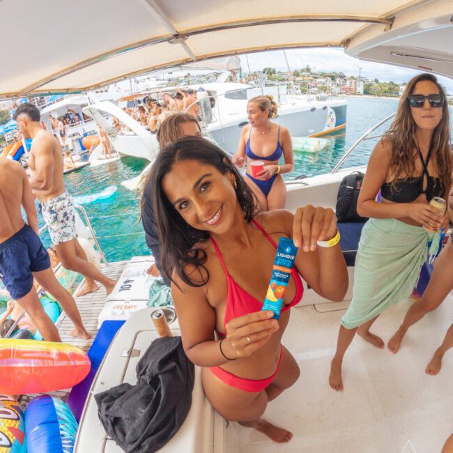 A smiling woman in a red bikini holds up a drink mix packet on a boat surrounded by people in swimwear enjoying a party, with boats and blue water in the background.