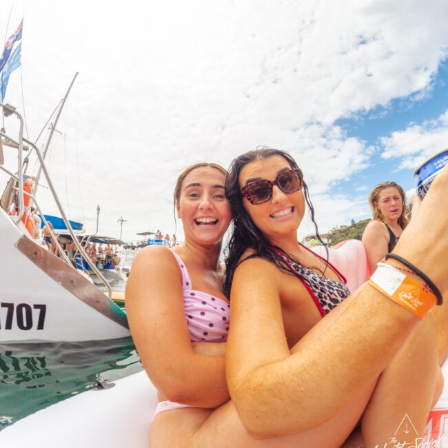 Two smiling women in swimsuits pose closely together on an inflatable float near a boat in the water, surrounded by other people. One wears sunglasses and both appear happy under a partly cloudy sky.