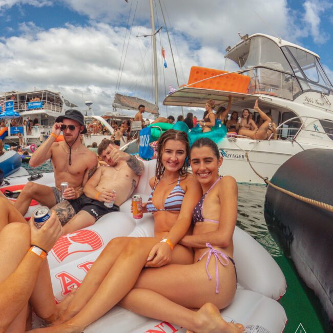 A group of young adults in swimsuits relax on inflatable rafts in the water, smiling and holding drinks, with boats and other people in the background under a partly cloudy sky.