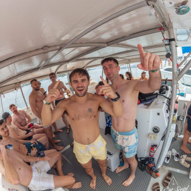 A group of young men in swimwear are smiling and pointing upwards while standing and sitting on a boat under a canopy, with the ocean visible in the background.
