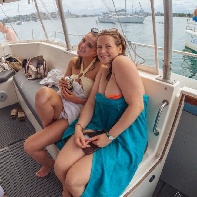 Two women in swimwear sit smiling on a boat bench, one holding a drink can and the other wrapped in a blue towel. Water, boats, and a distant city skyline are visible through the windows.