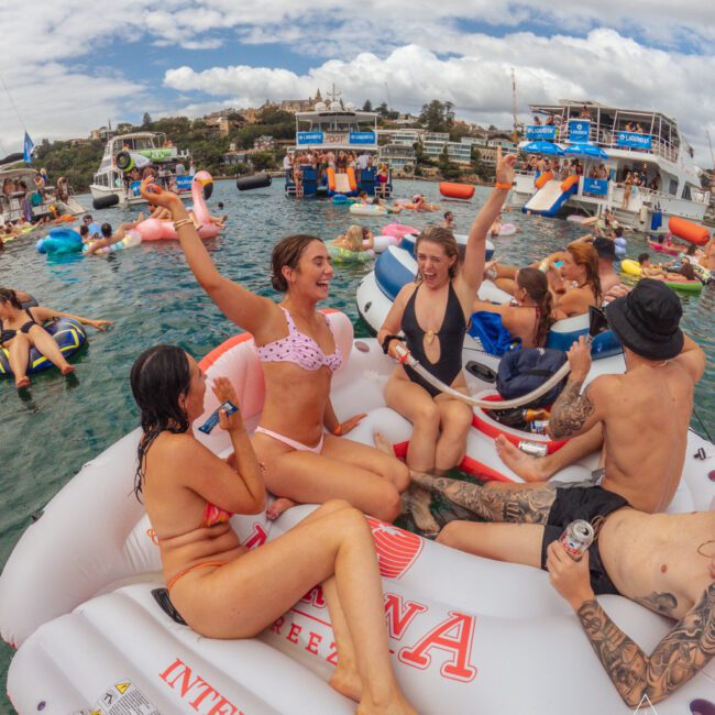 A group of young adults in swimsuits relax and cheer on a large inflatable float in the water, surrounded by others on floats and boats, under a partly cloudy sky at a lively outdoor party.