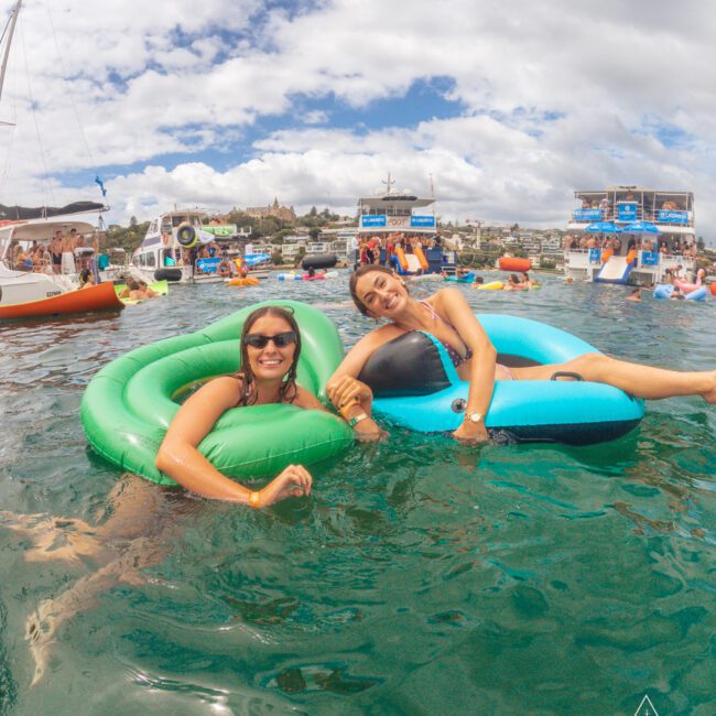 Two women in sunglasses smile while relaxing on green and blue pool floats in clear water, surrounded by other people, boats, and floating docks on a partly cloudy day.
