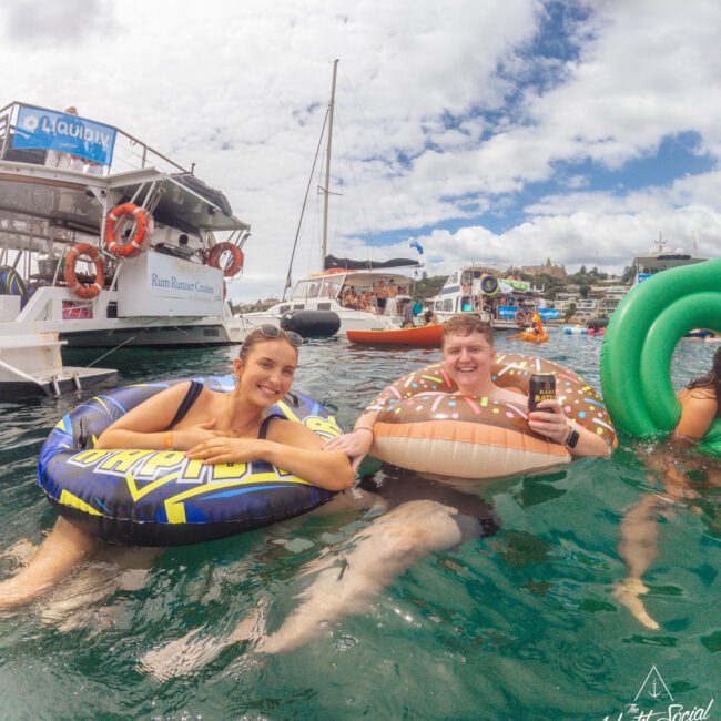Two people float in the ocean on inflatable tubes near several boats. They are smiling and holding drinks, enjoying a sunny day with others in the background. The scene is lively with blue skies and scattered clouds.
