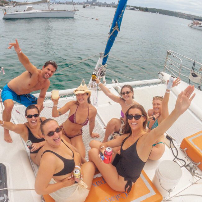 A group of six smiling people in swimsuits sit and raise drinks on a sailboat, with water, other boats, and a city skyline visible in the background. Everyone looks cheerful and is waving or posing for the camera.