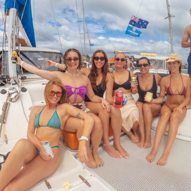 Six women in swimsuits sit and smile on a sailboat, holding drinks. The sky is partly cloudy, and Australian flags can be seen in the background. The group appears relaxed and cheerful, enjoying a day out on the water.