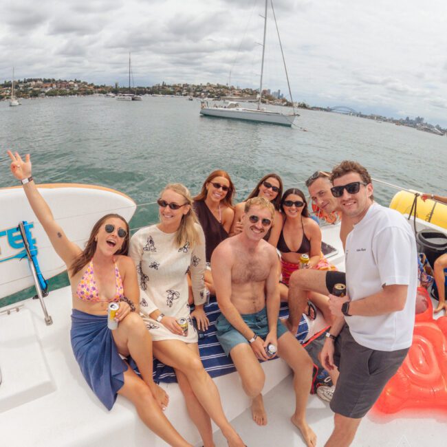A group of eight people are smiling and posing together on a boat, with water, a sailboat, and a cityscape in the background. It is daytime with cloudy skies, and everyone looks relaxed and cheerful.
