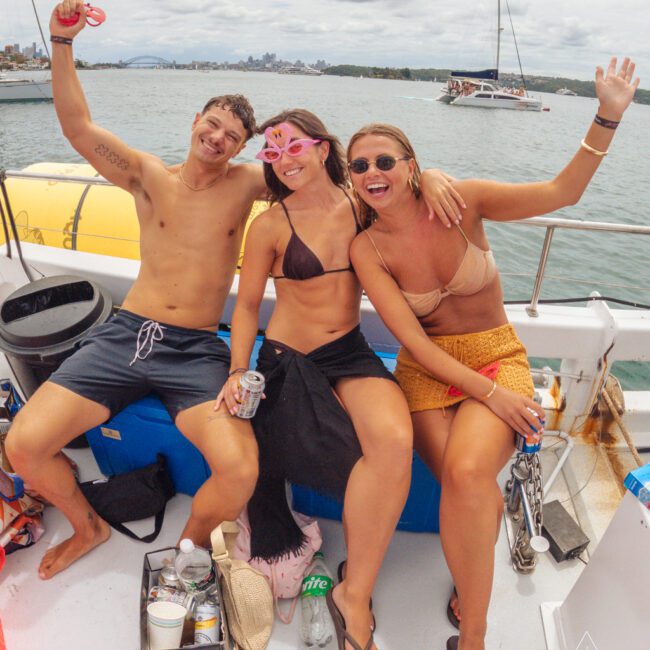 Three young adults in swimwear sit on a boat, smiling and posing for the camera. One woman wears pink novelty glasses, while the others hold drinks. The water and city skyline are visible in the background.