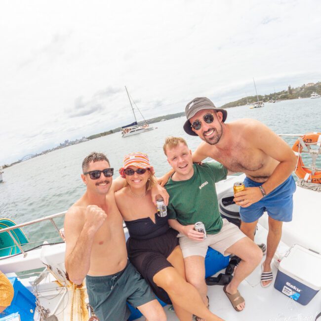 Four friends—three men and one woman—smile and pose together on a boat, holding drinks. The background shows calm water, more boats, and a cloudy sky. Everyone appears to be enjoying a casual day out.