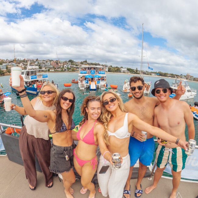 A group of six smiling young adults in swimwear pose together on a boat, holding drinks. Other boats and people are visible on the water behind them, under a cloudy sky.