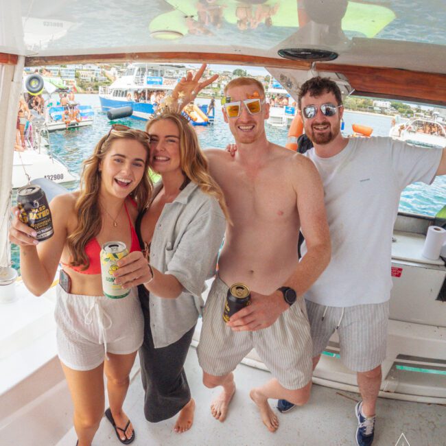 Four friends smile and pose with drinks on a boat during a sunny day, with other boats and people in the water visible in the background. One person gives a peace sign, and everyone appears to be enjoying a festive atmosphere.