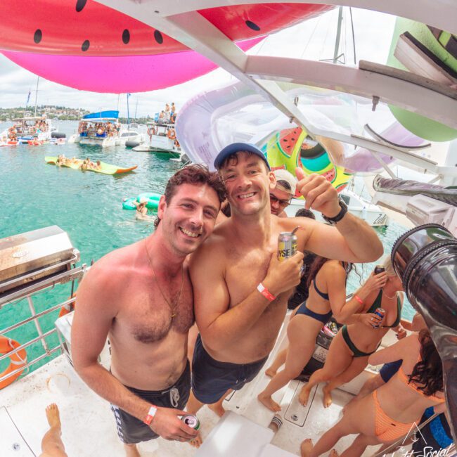 Two smiling shirtless men with drinks pose on a boat at a lively party with people in swimsuits. Inflatable watermelons and bright decorations hang above, and boats float on the turquoise water in the background.