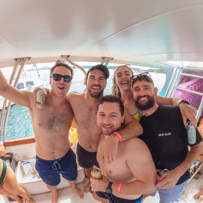 Five people in swimsuits pose smiling on a boat with drinks in hand. The background shows blue water and other boats. It appears to be a sunny, fun day, and everyone looks happy and relaxed.