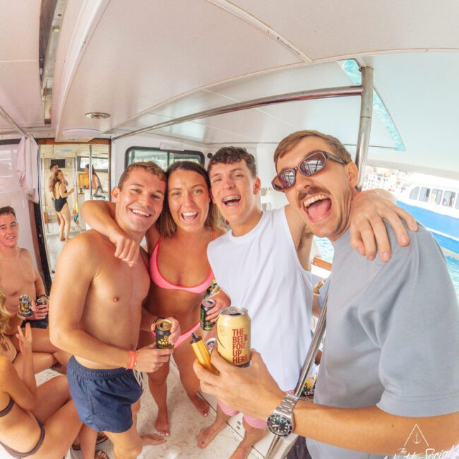 A group of smiling young adults in swimsuits and casual clothes hold drinks and pose together on a boat, with more people and water visible in the background.