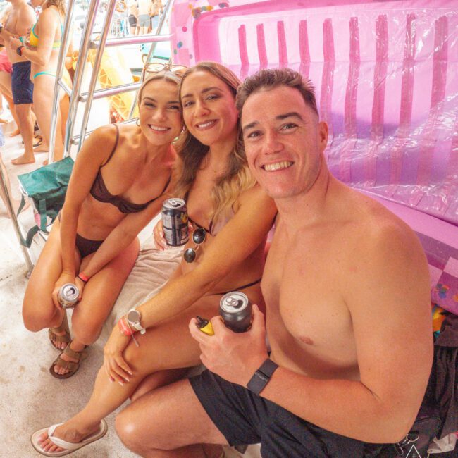 Three young adults in swimwear, smiling and holding cans, sit together by a large pink inflatable at a lively pool party. Other people mingle and lounge in the background.