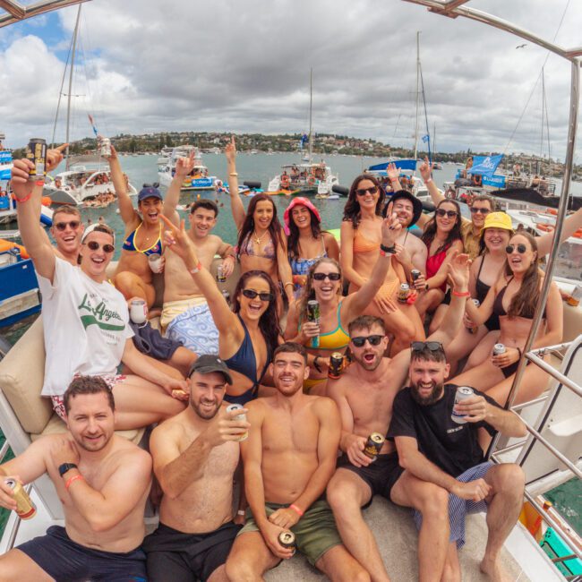 A large group of young adults in swimwear sit and stand on a boat, smiling, raising drinks, and cheering. Other boats and people are visible in the water behind them under a cloudy sky.