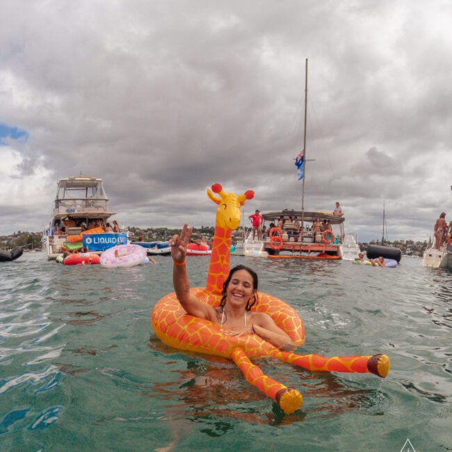 A smiling woman in an inflatable giraffe float raises a peace sign while floating on water, with boats and people in the background under a cloudy sky.