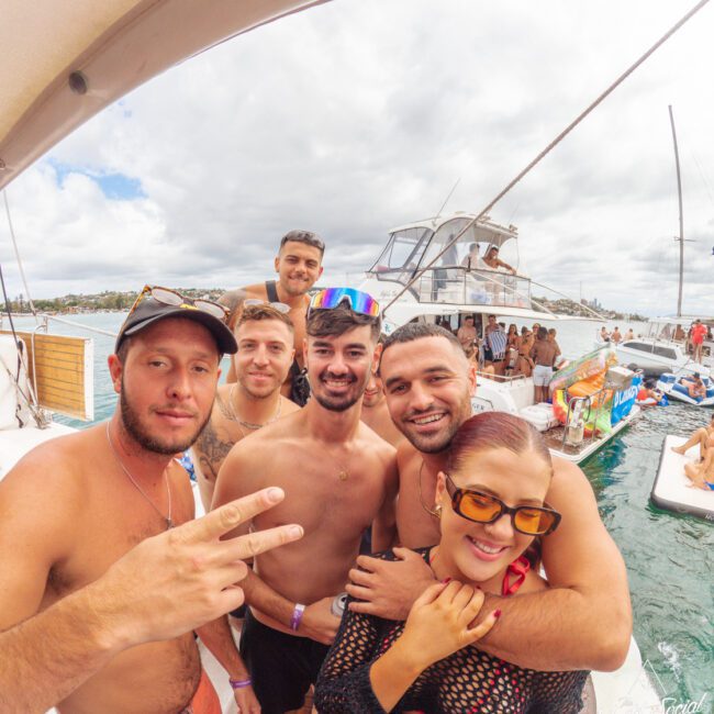 A group of six young adults smile and pose for a selfie on a boat, with water, other boats, and people in the background under a cloudy sky. One person in front flashes a peace sign.