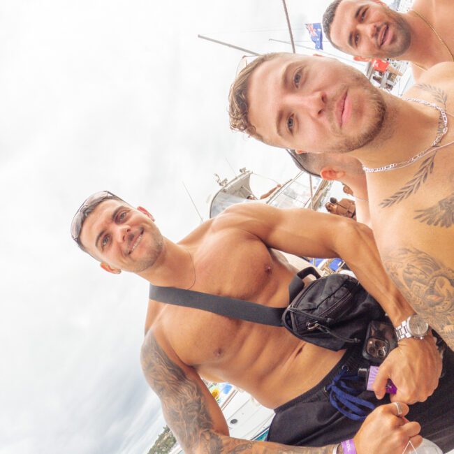 Three young men with tattoos pose shirtless on a docked boat, smiling at the camera. The background shows other boats, water, and a cloudy sky, suggesting a lively, beachside or yacht party atmosphere.