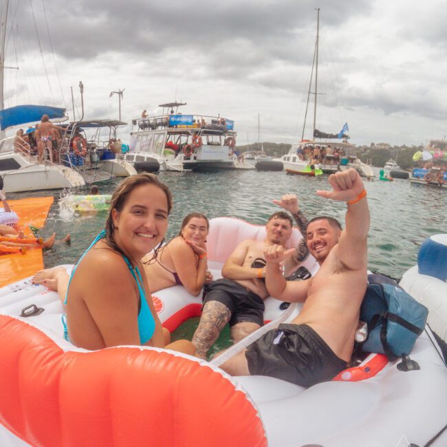 Four people relax and smile on an inflatable float in the water, surrounded by boats and other people. It’s a cloudy day, and the group looks happy and festive at a boating event.