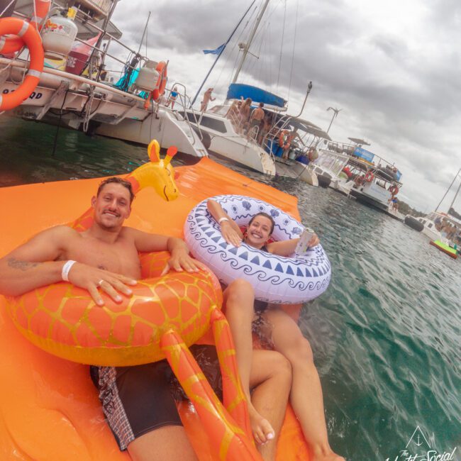 Two people relax and smile on colorful inflatable floats in the water near boats during a cloudy day, with others socializing and enjoying themselves on the nearby boats.