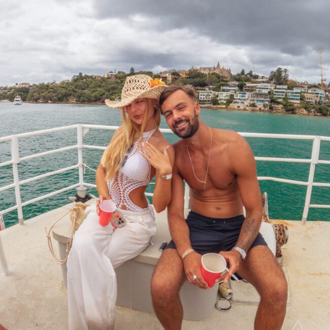 A woman in a crocheted hat and white outfit stands next to a shirtless man in swim trunks; both are holding red cups and smiling on a boat, with water, houses, and a cloudy sky in the background.
