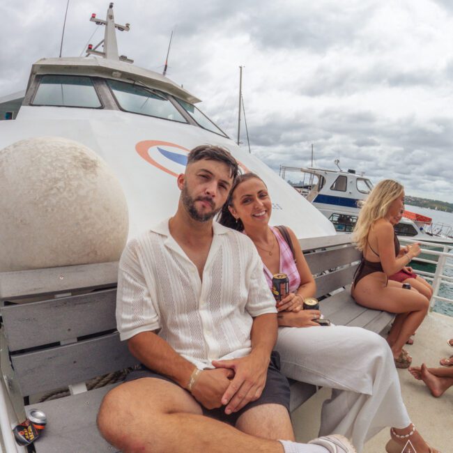 A man and woman sit closely together on a yacht bench, smiling at the camera. The man wears a white shirt and shorts, and the woman wears a sleeveless top. Other people relax in the background. Cloudy sky overhead.