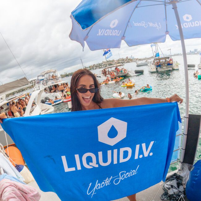 A smiling woman in sunglasses holds a blue "LIQUID I.V. Yacht Social" towel on a boat, with people swimming and socializing in the water behind her under cloudy skies.