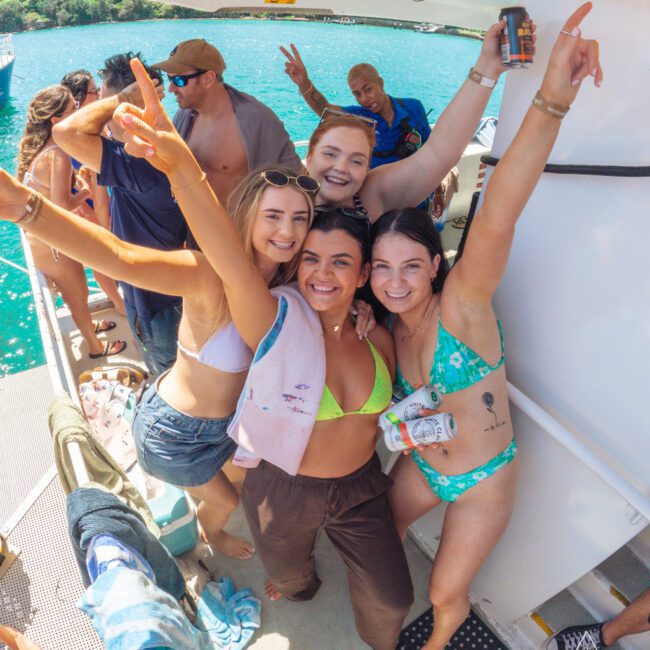 Four women in swimsuits smile and pose with raised arms on a boat, surrounded by other people enjoying a sunny day on the water. The turquoise sea and lush green coastline are visible in the background.