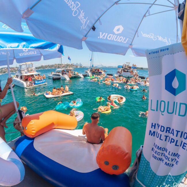 People relax on colorful inflatables in a crowded, sunny harbor. The scene is viewed from under branded blue and white umbrellas beside a floating hydration drink station. Boats and city skyline are visible in the background.