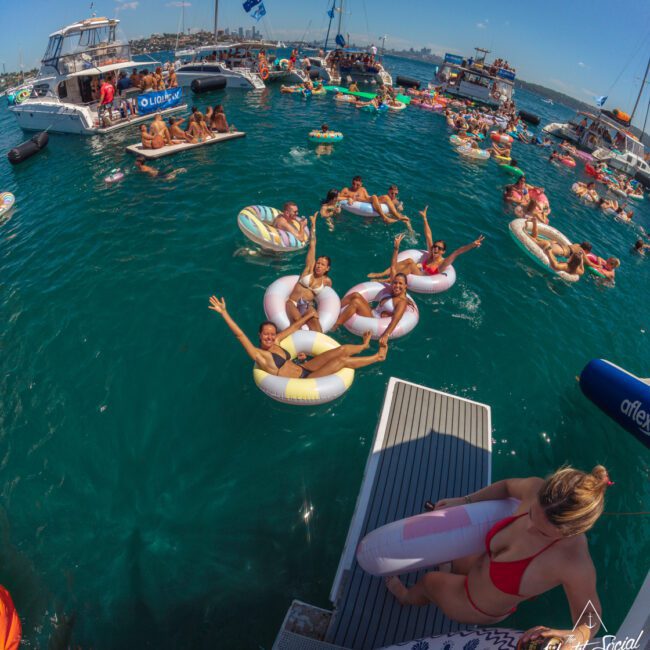 A lively scene of people relaxing on colorful inflatables and boats in bright blue water under a sunny sky. Several friends smile and wave at the camera, enjoying a festive, social day on the water.