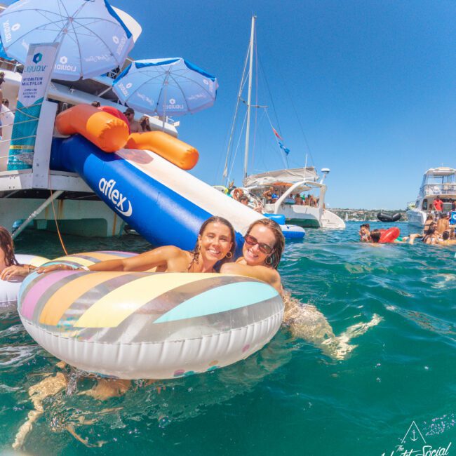 Two women smiling and relaxing on a colorful float in the ocean beside a yacht with a water slide. Other people, boats, and umbrellas are visible in the background under a clear blue sky.