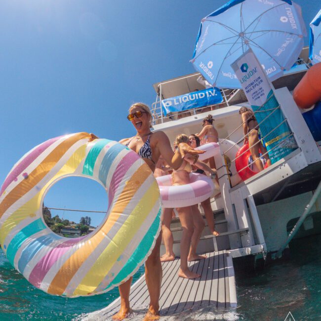 A group of people in swimsuits stand on a boat’s platform, holding colorful inflatable rings under bright sun and blue sky. Some are shaded by branded umbrellas, with water and shoreline visible in the background.