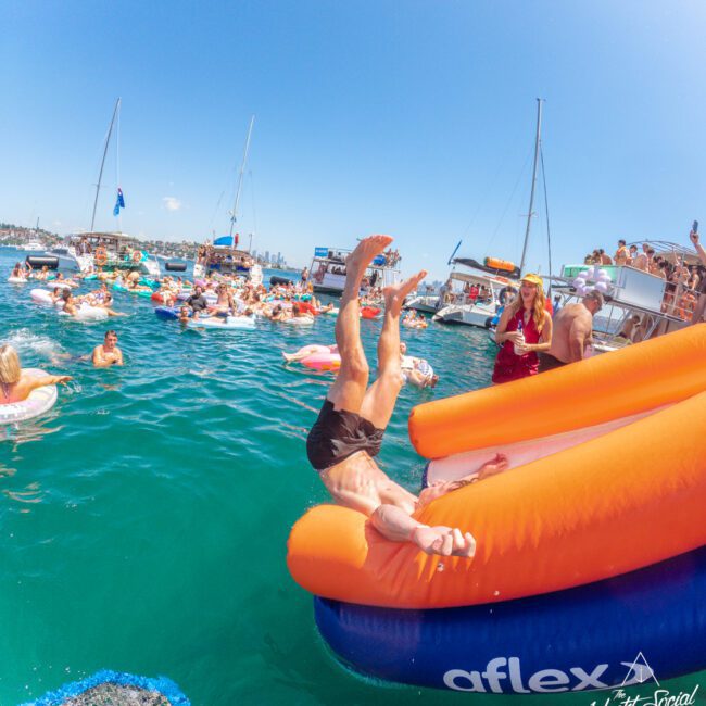 A man slides headfirst off an orange and blue inflatable slide into the water, surrounded by boats and people enjoying a sunny day at a lively boat party. The water is bright blue and the sky is clear.