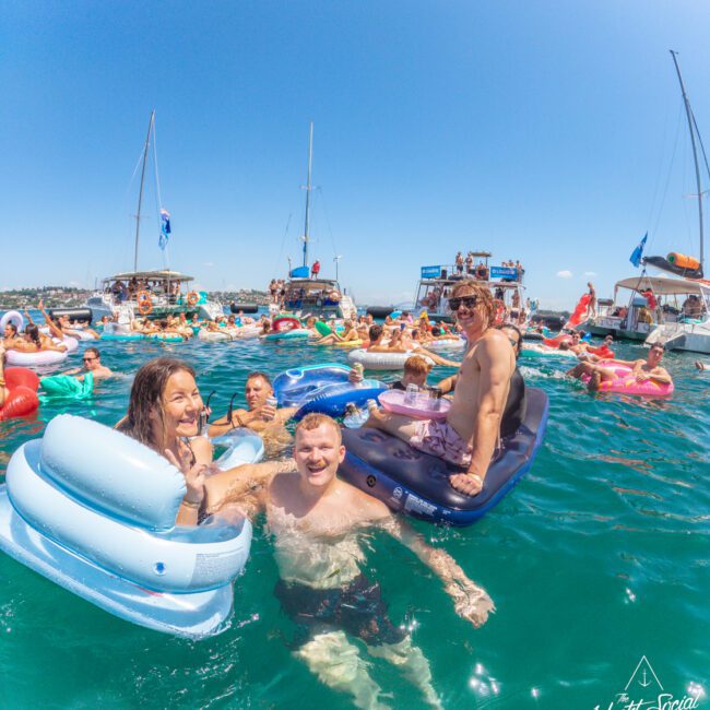 A group of people smiling and relaxing on inflatable floats in the water near anchored boats at a lively yacht party under clear blue skies. The mood is festive and joyful.