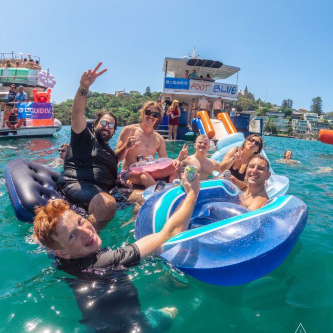 A group of smiling people relax on inflatables in clear blue water near a party boat, enjoying drinks and making peace signs on a sunny day. Other boats and a slide are visible in the background.