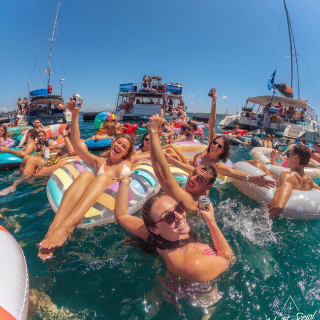 A group of people relax on colorful pool floats in the ocean, holding drinks and smiling, with boats and more partygoers in the background under a clear blue sky.