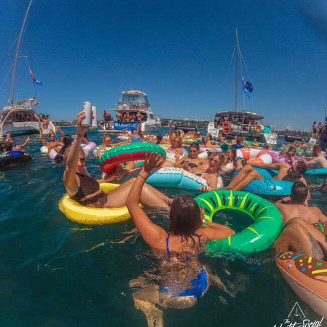 A group of people relax and have fun on colorful inflatable floaties in the ocean near several anchored boats under a clear blue sky. The scene is lively and festive, with people raising drinks and smiling.