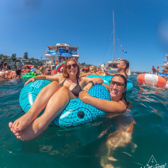 Two women in swimsuits smile and relax on a blue inflatable float in the water with other people and boats in the background on a sunny day. The scene is lively and festive. A "Yacht Social Club" logo is in the corner.