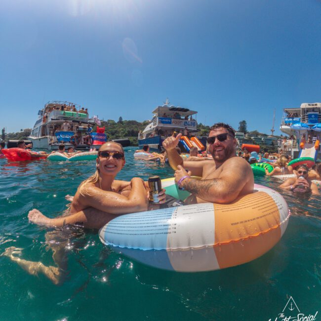 Two people smile and hold drinks while relaxing on an inflatable float in the water, surrounded by others and boats, during a sunny day at a social event. The Yacht Social Club logo is visible in the corner.