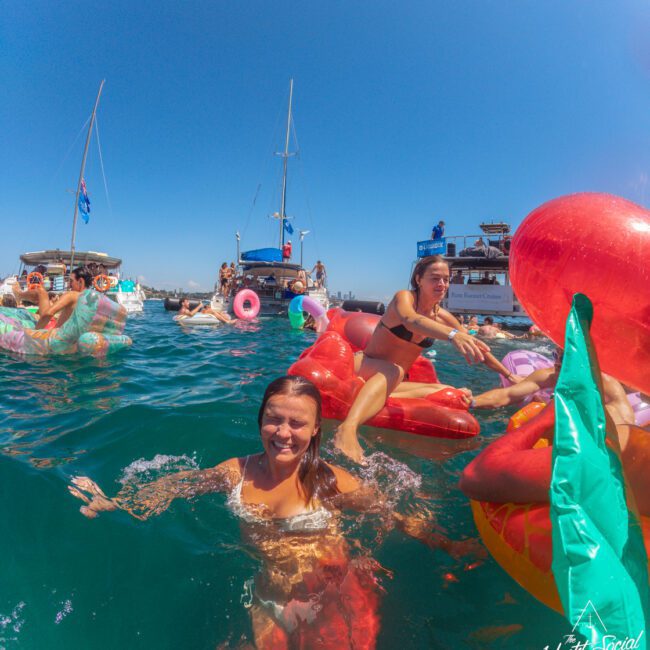 A woman smiles while swimming in clear blue water near inflatable pool floats and boats. Other people relax on colorful inflatables in the background under a sunny sky. The Yacht Social Club logo is in the bottom right corner.