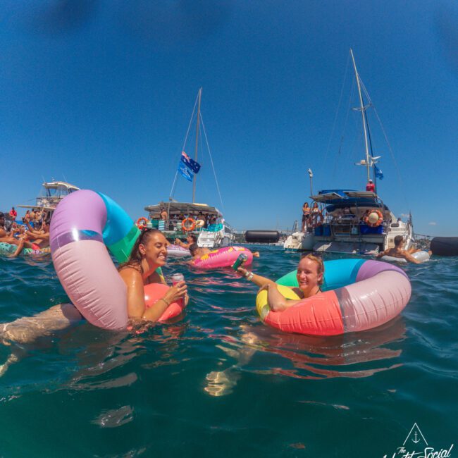 Two women float on colorful inflatable rings in clear blue water near several boats, with others swimming and relaxing in the background under a sunny sky. The scene has a festive, social atmosphere.