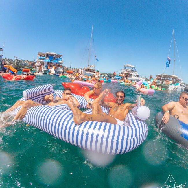 A group of people relax on striped inflatable rafts in a lively, crowded party on the water, surrounded by boats and other colorful floaties under a clear blue sky.