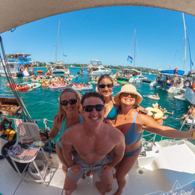 Four friends in swimsuits smile at the camera on a boat, surrounded by other boats and people floating in the water at a lively outdoor gathering under a sunny blue sky.