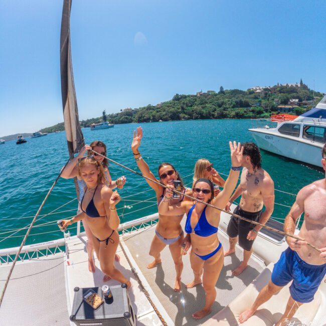 A group of people in swimsuits smile and wave while standing on a boat deck with snacks and drinks; the blue ocean and another boat are visible in the background under a clear sky.