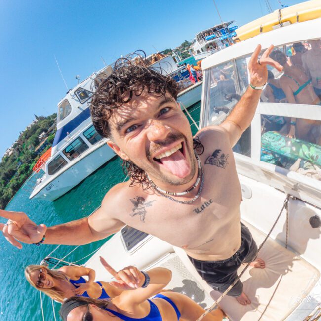 A man with curly hair sticks out his tongue and flashes peace signs on a boat, surrounded by friends in swimsuits. Other boats and clear blue water are visible in the background under a bright sky.