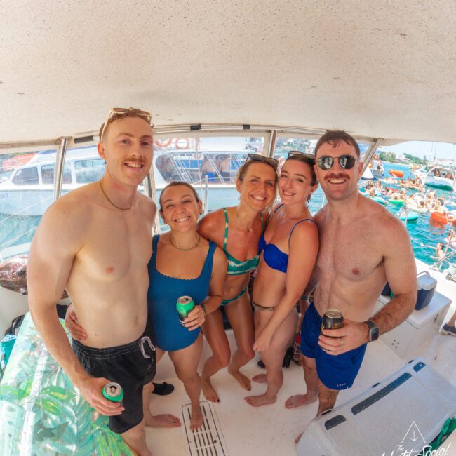 Five people in swimwear smiling and posing together on a boat, holding canned drinks. Other boats and people are visible in the water in the background. The atmosphere appears sunny and fun.