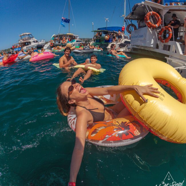A group of people enjoy a lively party on and around boats, floating on inflatable rings in clear blue water; a smiling woman in sunglasses reaches for the camera. The scene is sunny and festive.