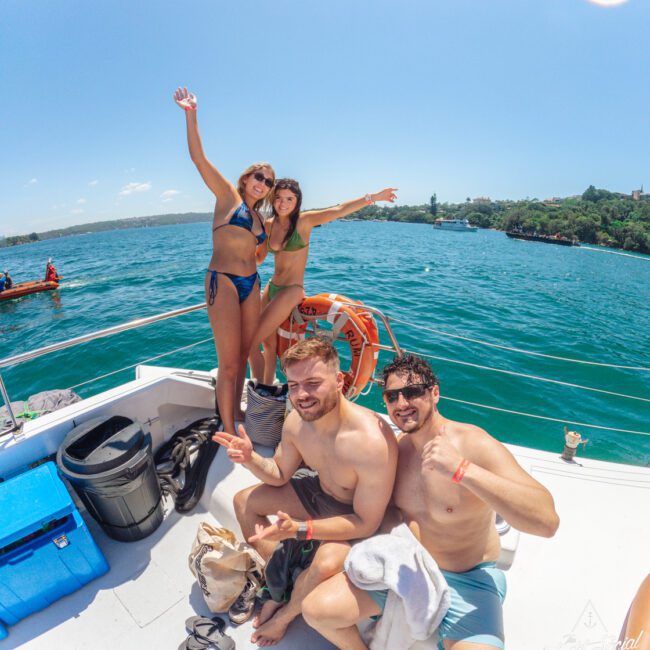Four young adults in swimsuits pose and smile on a boat under a bright blue sky, with two women standing and two men sitting, surrounded by clear turquoise water and distant greenery.